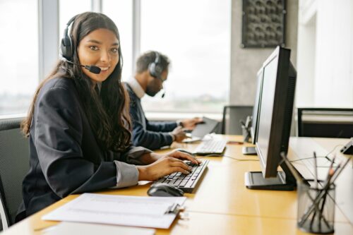 female Call Center Help Desk Representative looks into camera with computer screen to right and man in background