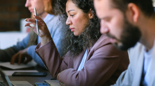 woman in middle distance looks at computer and holds pen, man in foreground and background