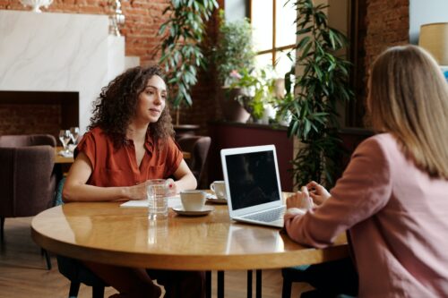 woman with dark curly hair being interviewed by another woman wearing pink in a room with plants and a brick wall