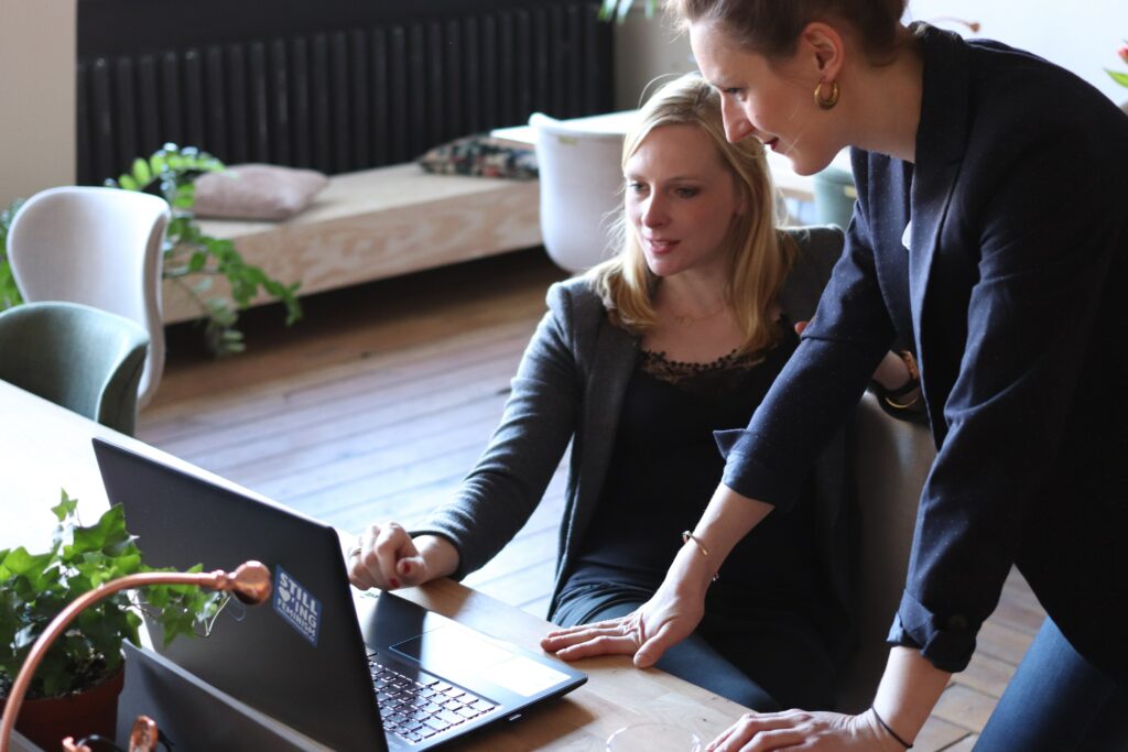 two women looking at a laptop, one standing, one sitting.