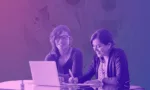 Two women co-working at a laptop on a conference table, one writing notes, both smiling.
