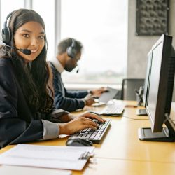 female Call Center Help Desk Representative looks into camera with computer screen to right and man in background