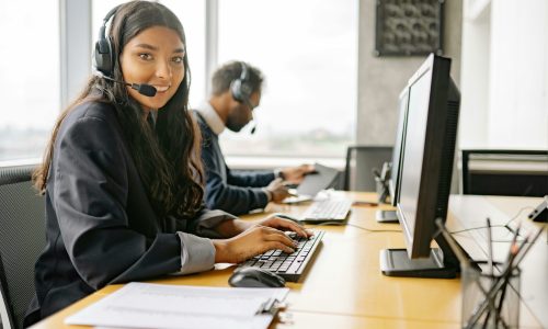 female Call Center Help Desk Representative looks into camera with computer screen to right and man in background