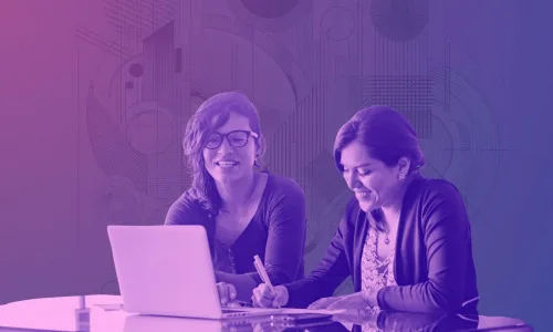Two women co-working at a laptop on a conference table, one writing notes, both smiling.