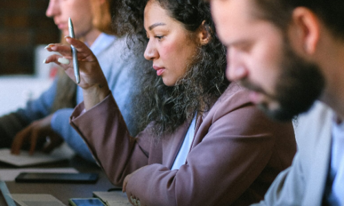 woman in middle distance looks at computer and holds pen, man in foreground and background