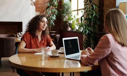 woman with dark curly hair being interviewed by another woman wearing pink in a room with plants and a brick wall